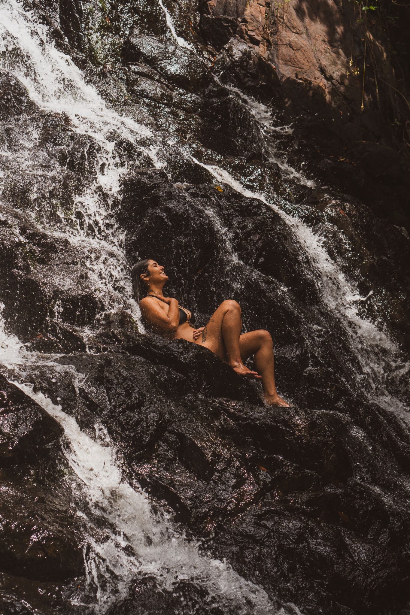 A person sits on dark rocks beneath flowing water.