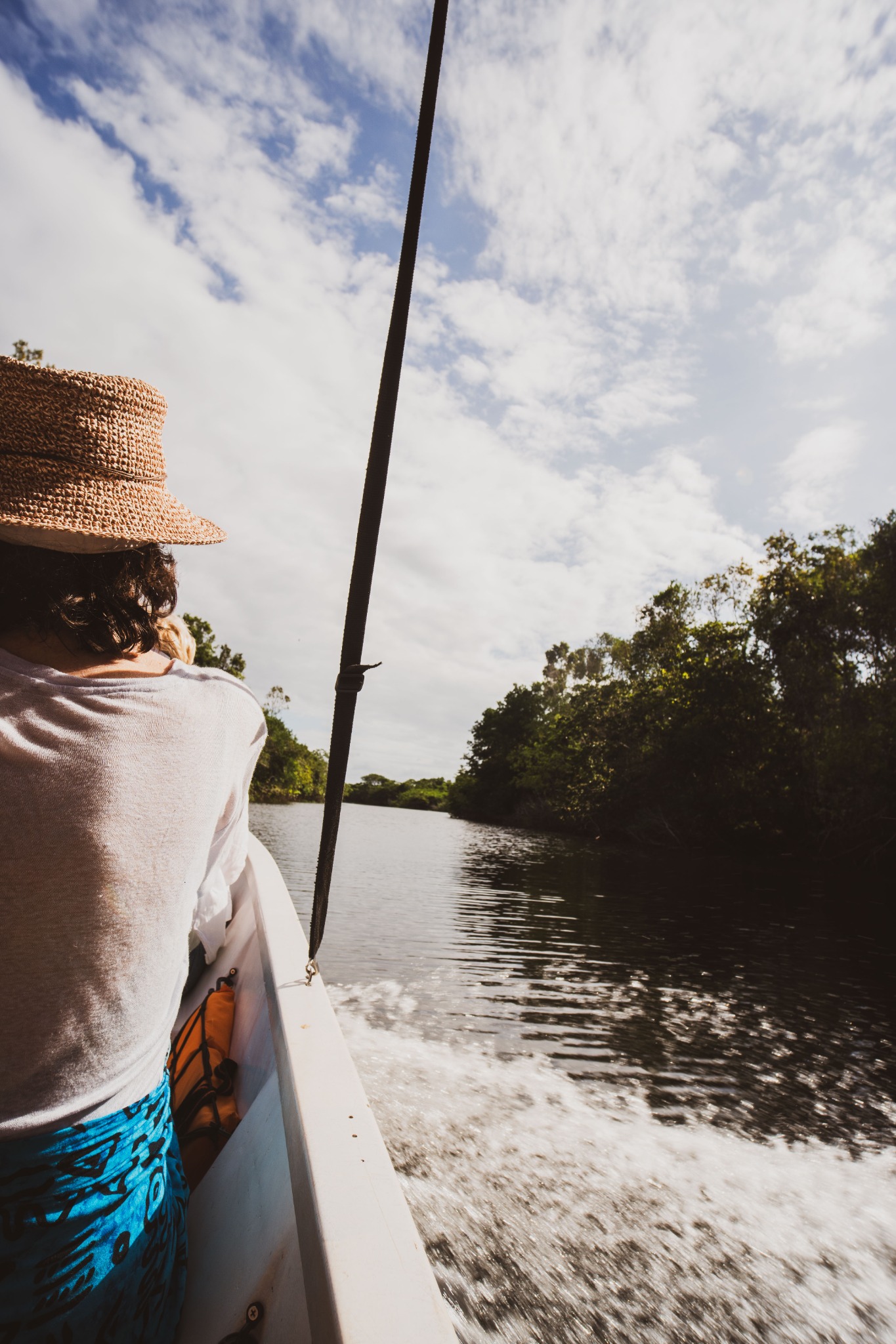 A passenger in a straw hat looks ahead from a moving boat on tree-lined water.