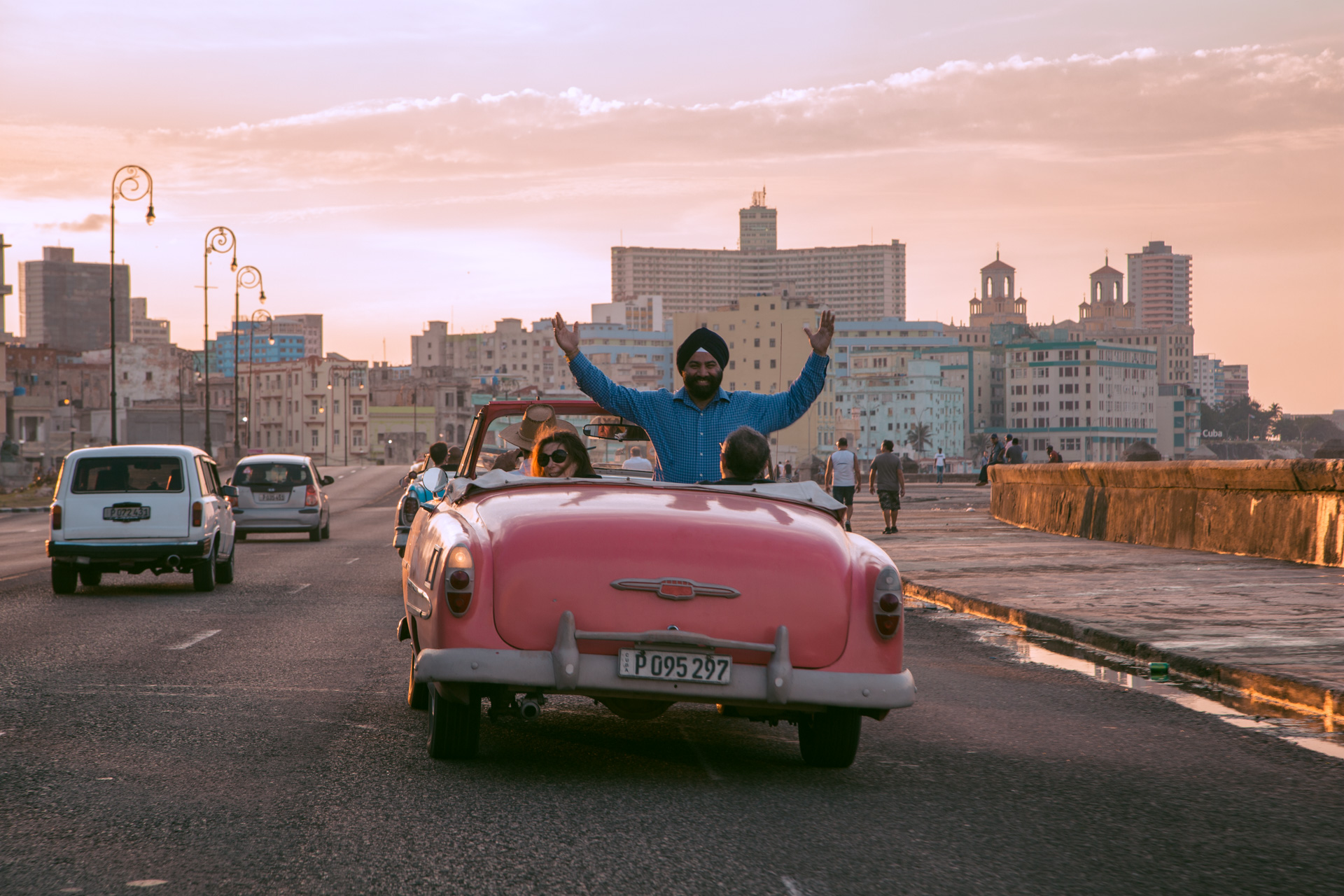 Navneet raises his arms from a pink vintage convertible on Havana’s Malecón at sunset.